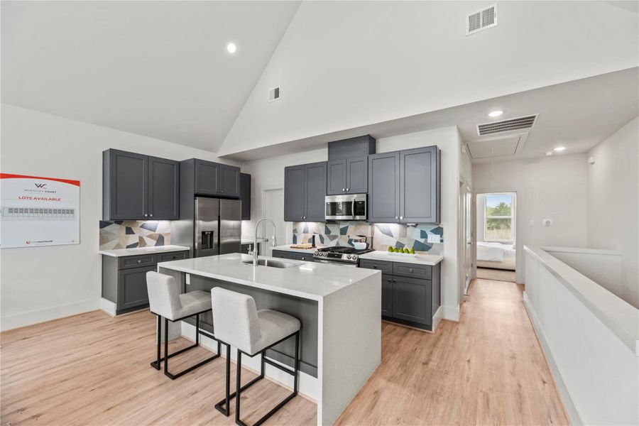Step into a modern kitchen featuring sleek gray cabinets, a spacious island with seating, stainless steel appliances, and a chic backsplash. Light wood floors and an open layout make the space feel bright, airy, and perfect for everyday living or entertaining.