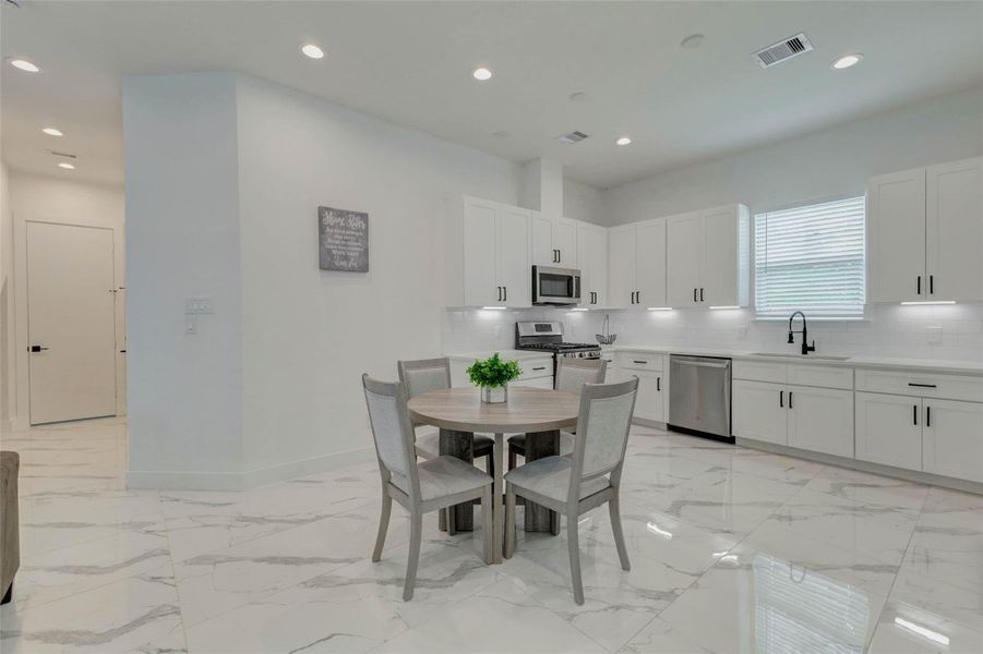 Spacious dining and kitchen area with glossy marble-look tile floors, a round dining table, white cabinetry, black hardware, stainless appliances, under-cabinet lighting, and a bright window over the sink. The open layout feels fresh, functional, and perfect for casual meals.