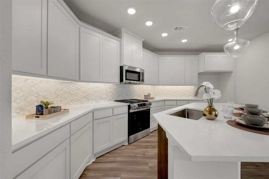 Kitchen featuring white shaker-style cabinetry, white countertops, textured tile backsplash with under-cabinet lighting, stainless steel appliances, and a central island with an undermount sink