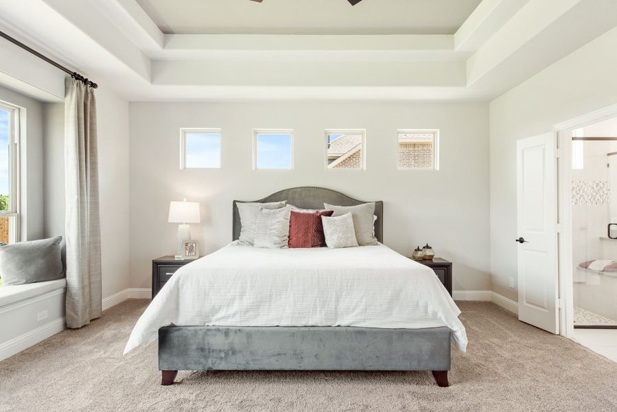 Master bedroom with tray ceiling, gray upholstered bed, carpet flooring, and ensuite bathroom access Master bedroom with tray ceiling, gray upholstered bed, carpet flooring, and ensuite bathroom access