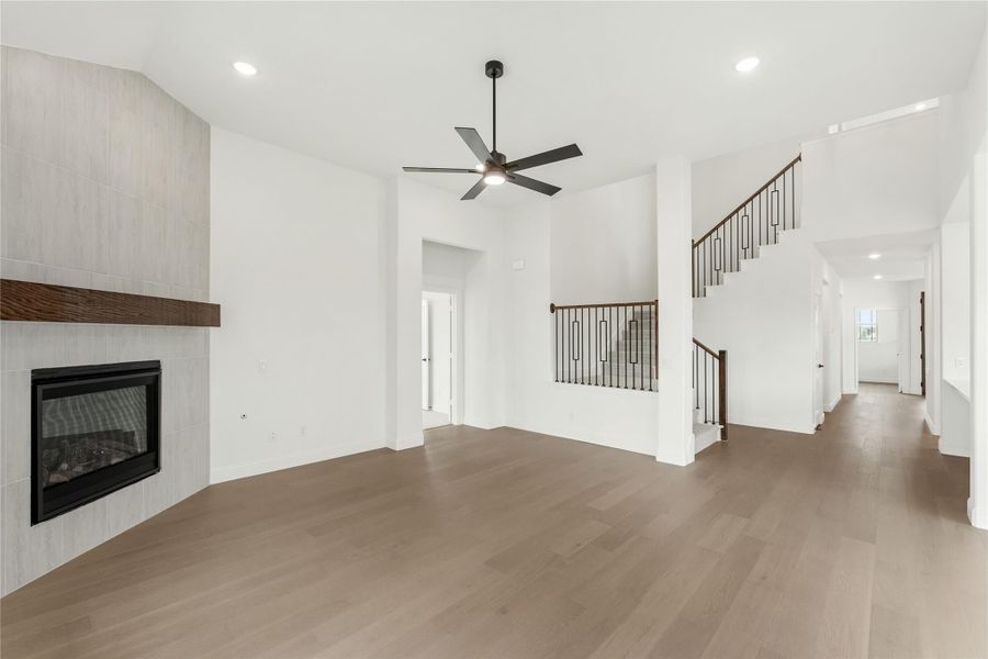 Open living room with tile fireplace, ceiling fan, hardwood floors, and staircase visible in background. Open living room with tile fireplace, ceiling fan, hardwood floors, and staircase visible in background.