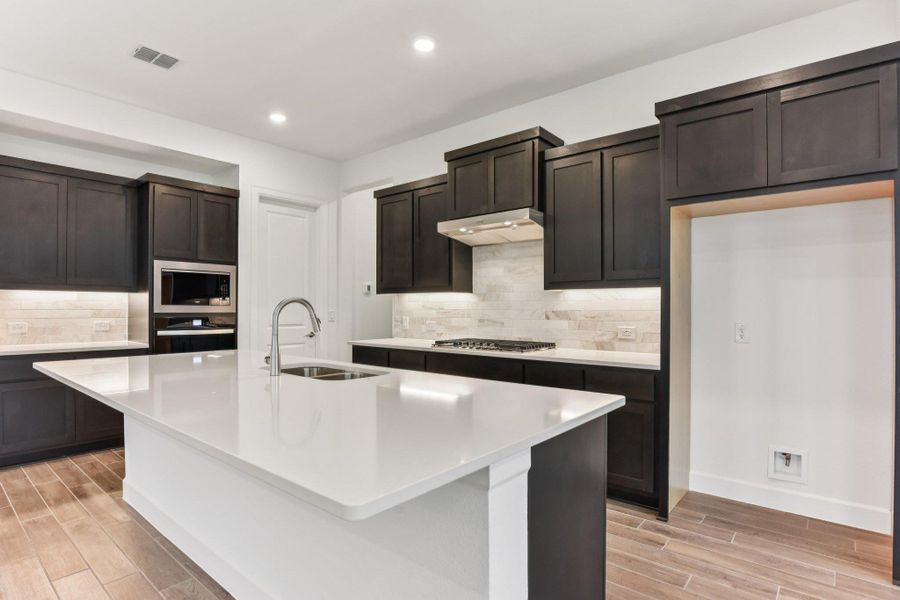 Kitchen featuring wood finish floors, light stone counters, appliances with stainless steel finishes, backsplash, and dark brown cabinetry Kitchen featuring wood finish floors, light stone counters, appliances with stainless steel finishes, backsplash, and dark brown cabinetry