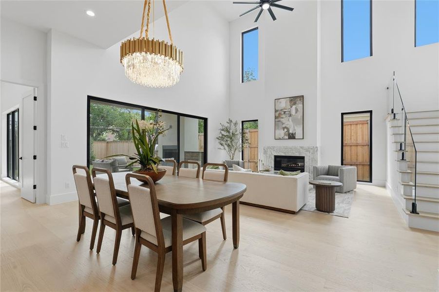 Open-concept living space featuring wood-finish flooring, a contemporary chandelier, a fireplace with a stone surround, black-framed sliding glass doors, and a staircase with glass panel railing Open-concept living space featuring wood-finish flooring, a contemporary chandelier, a fireplace with a stone surround, black-framed sliding glass doors, and a staircase with glass panel railing