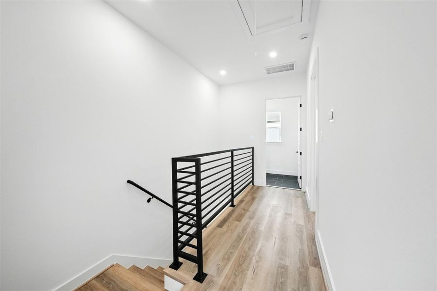 Third-floor hallway with sleek railing and wide-plank flooring, leading to the primary bedroom on the right and a utility room straight ahead. Clean lines and recessed lighting create a bright, modern transition space. Third-floor hallway with sleek railing and wide-plank flooring, leading to the primary bedroom on the right and a utility room straight ahead. Clean lines and recessed lighting create a bright, modern transition space.