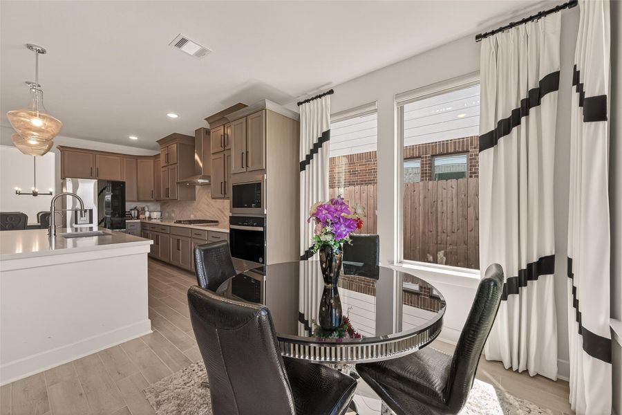 Modern kitchen and dining area featuring sleek cabinetry, stainless steel appliances, and a large island with pendant lighting. The space is bright with natural light from large windows with stylish curtains, and includes a cozy dining table, creating an inviting atmosphere.