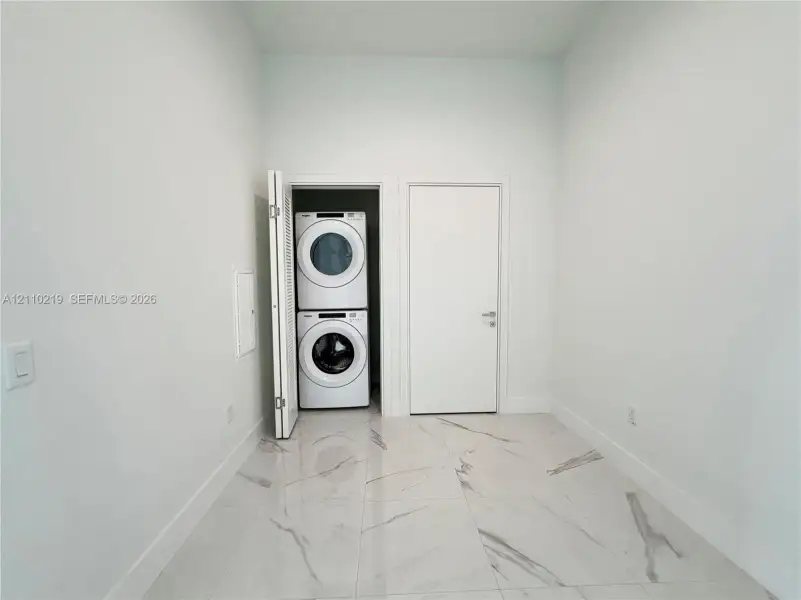 Utility area featuring large-format tile flooring with a neutral veined pattern, crisp white walls, and a recessed closet housing a stacked washer and dryer unit