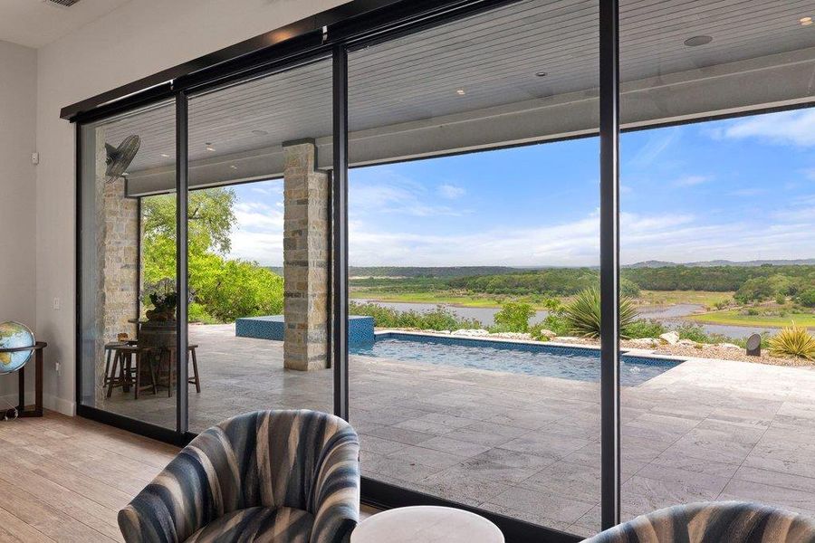 Large format sliding glass doors opening to an outdoor living area with a stone patio, an infinity-edge pool, and a panoramic lake and hill country landscape