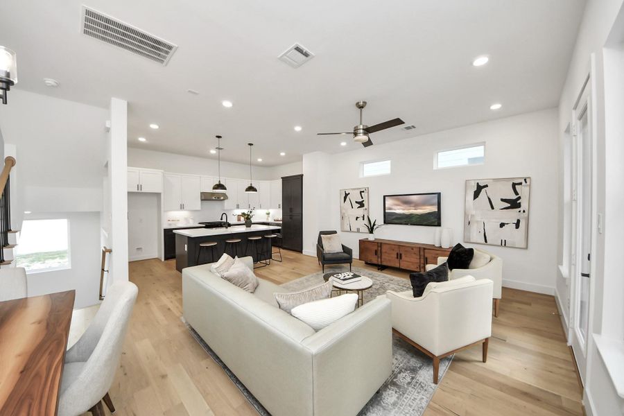 This photo showcases a modern, open-concept living area with a kitchen and living room. The space features light wood flooring, white walls, recessed lighting, and stylish furnishings. The kitchen has sleek black accents, an island with seating, and pendant lighting. The living room includes comfortable seating, a ceiling fan, and contemporary artwork. This photo showcases a modern, open-concept living area with a kitchen and living room. The space features light wood flooring, white walls, recessed lighting, and stylish furnishings. The kitchen has sleek black accents, an island with seating, and pendant lighting. The living room includes comfortable seating, a ceiling fan, and contemporary artwork.