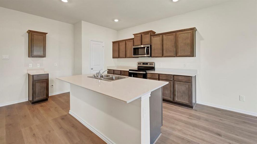 Kitchen featuring wood-finish flooring, dark wood cabinetry, light-toned countertops, a central island with an undermount double sink, and stainless steel appliances including a microwave and range