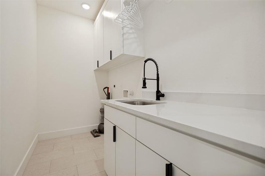 Laundry area featuring a built-in sink with a matte black gooseneck faucet, white cabinetry with black hardware, light-toned countertop, recessed ceiling lighting, and tiled flooring Laundry area featuring a built-in sink with a matte black gooseneck faucet, white cabinetry with black hardware, light-toned countertop, recessed ceiling lighting, and tiled flooring