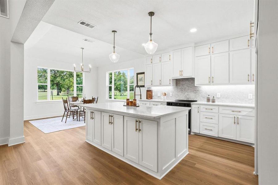 Open-concept kitchen featuring an expansive center island, white cabinetry with gold-tone hardware, a tiled backsplash, wood-finish flooring, and recessed lighting Open-concept kitchen featuring an expansive center island, white cabinetry with gold-tone hardware, a tiled backsplash, wood-finish flooring, and recessed lighting
