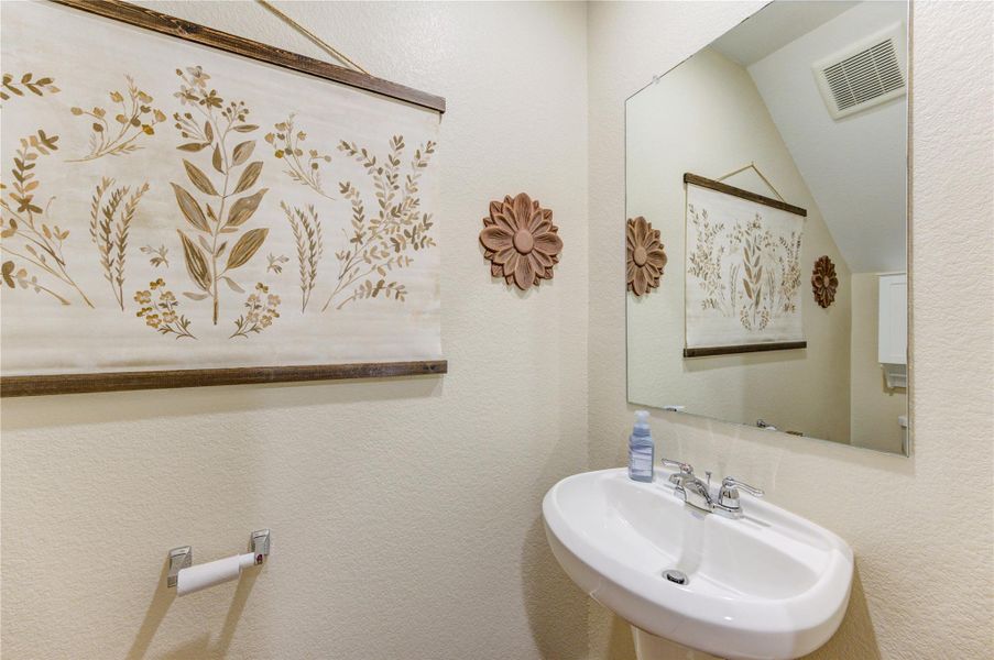 This is a cozy half-bathroom featuring a white pedestal sink, a large mirror, and tasteful wall decor with floral and wooden accents. The neutral color scheme creates a clean and inviting atmosphere.