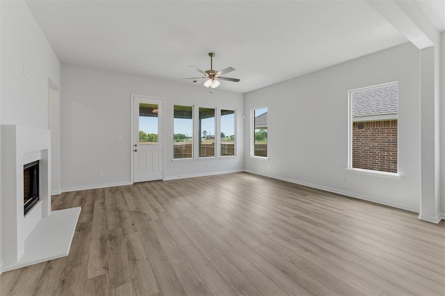 Spacious living area featuring light wood-finish flooring, a contemporary fireplace with a white surround, and multiple windows providing ample natural light Spacious living area featuring light wood-finish flooring, a contemporary fireplace with a white surround, and multiple windows providing ample natural light