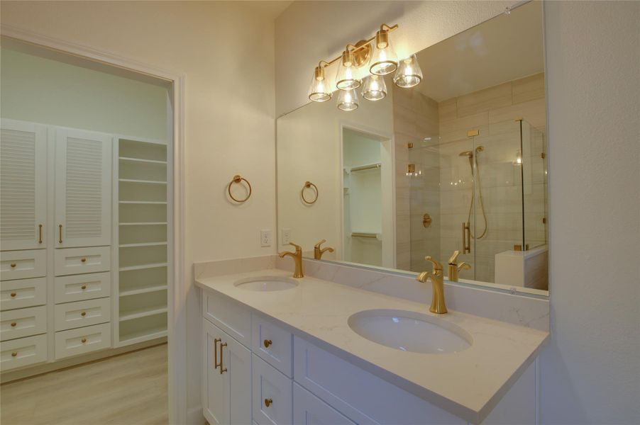 Primary bath with a dual-sink quartz vanity, goldfixtures, and designer lighting. Frameless glassshower nearby. Direct access to a spacious walk-incloset with custom built-ins and open shelving.Warm neutral finishes and wide-plank floorscomplete the look. Primary bath with a dual-sink quartz vanity, goldfixtures, and designer lighting. Frameless glassshower nearby. Direct access to a spacious walk-incloset with custom built-ins and open shelving.Warm neutral finishes and wide-plank floorscomplete the look.