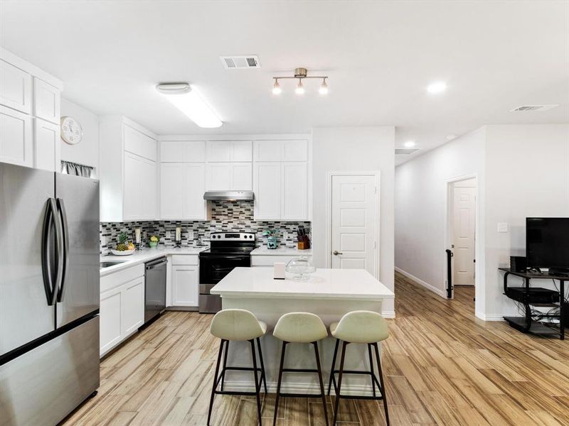 Open-concept kitchen featuring white cabinetry, a mosaic tile backsplash, a center island with countertop seating, stainless steel appliances, and wood-finish flooring