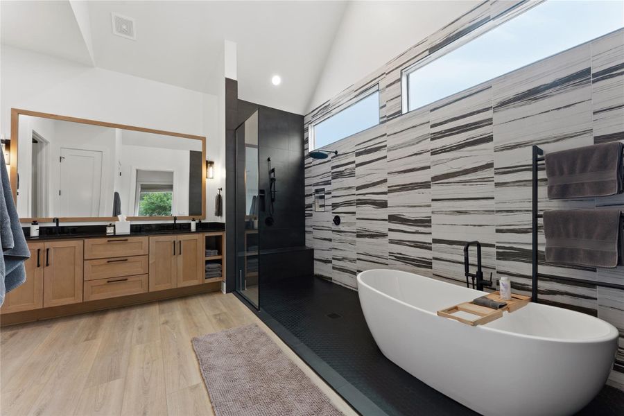 Contemporary bathroom featuring a freestanding soaking tub, a spacious glass-enclosed shower, dual vanities with wood cabinetry and dark countertops, large framed mirror, and light wood-finish flooring Contemporary bathroom featuring a freestanding soaking tub, a spacious glass-enclosed shower, dual vanities with wood cabinetry and dark countertops, large framed mirror, and light wood-finish flooring