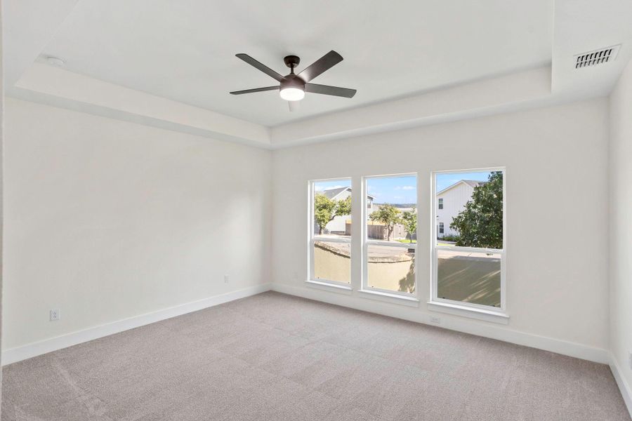 Spacious room featuring a tray ceiling, ceiling fan with integrated lighting, three large windows, gray carpeting, and white baseboards Spacious room featuring a tray ceiling, ceiling fan with integrated lighting, three large windows, gray carpeting, and white baseboards