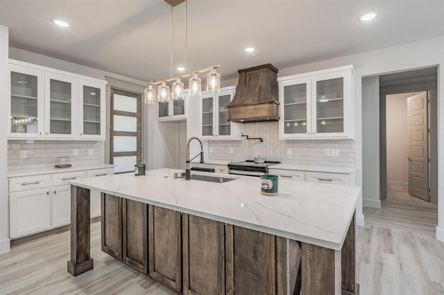 Kitchen with glass insert cabinets, white cabinets, tasteful backsplash, a center island with sink, and premium range hood