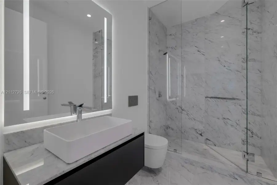 Floating vanity with integrated countertop, rectangular vessel sink, wall-to-wall mirror with vertical integrated lighting, glass-enclosed shower, and stone-finish wall and floor tiling