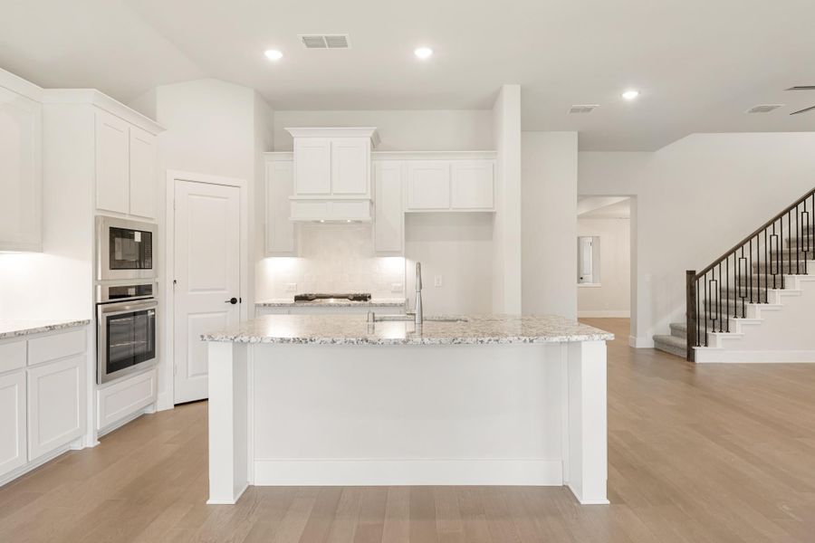 Open kitchen with white cabinets, granite island, stainless steel appliances, and light hardwood floors open to staircase area.