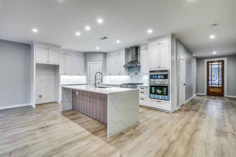 Kitchen with white cabinetry, sink, wall chimney range hood, a kitchen island with sink, and light wood-type flooring