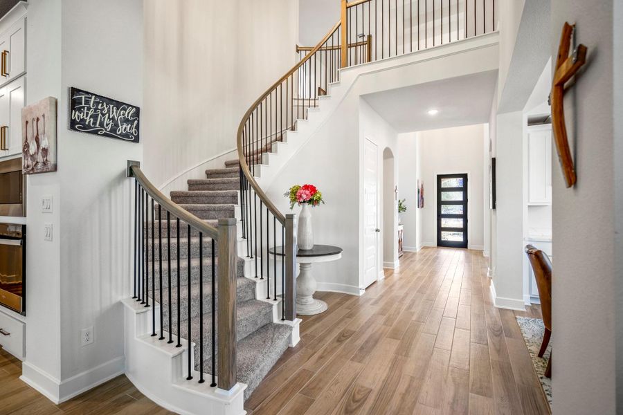 This inviting foyer features a beautiful, curved staircase with wrought iron railings. The space is bright and airy with wood flooring. A modern, black-framed door adds a stylish touch, and decorative accents enhance the welcoming ambiance.