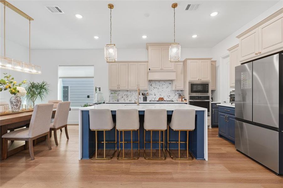 Open-concept kitchen featuring an oversized island with seating, stainless steel appliances, light-toned upper cabinetry, contrasting navy base cabinetry, and wood-finish flooring Open-concept kitchen featuring an oversized island with seating, stainless steel appliances, light-toned upper cabinetry, contrasting navy base cabinetry, and wood-finish flooring