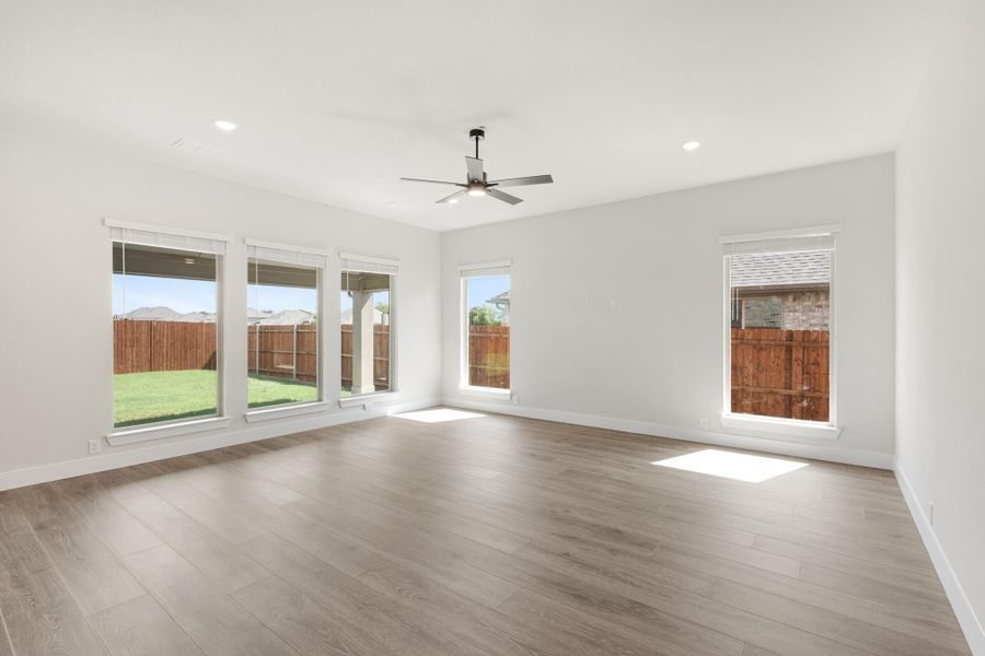 Empty living room with light wood floors, ceiling fan, and large windows overlooking a fenced backyard