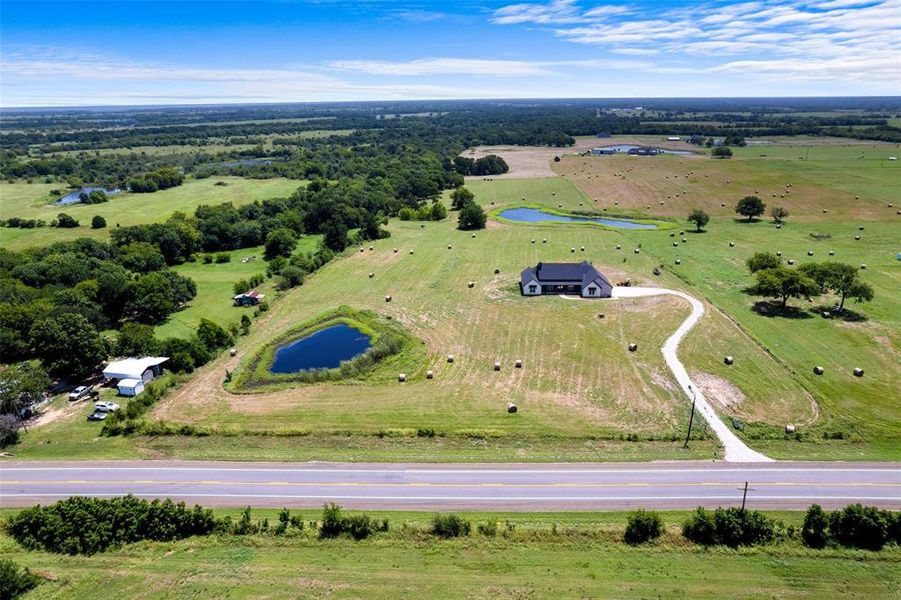 Expansive rural property featuring a modern single-story residence with a dark roof, a private pond, mature trees, and a winding driveway