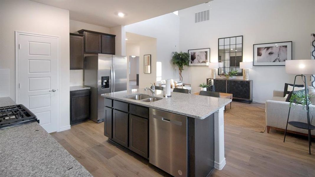 Kitchen featuring granite countertops, wood-finish flooring, stainless steel appliances, dark cabinetry, and a central island with an undermount double sink Kitchen featuring granite countertops, wood-finish flooring, stainless steel appliances, dark cabinetry, and a central island with an undermount double sink