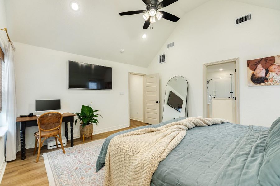 This bedroom features a vaulted ceiling with a ceiling fan, a mounted TV, and a cozy workspace by the window. It has space for a large bed, a full-length mirror, and an en-suite bathroom. The space is bright and welcoming, with modern decor and wood look flooring.