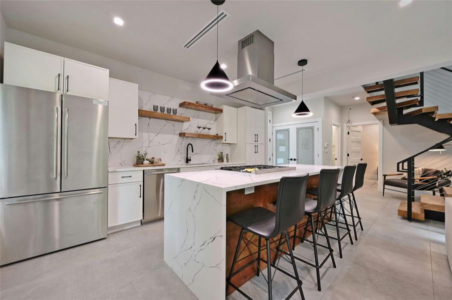 Stunning modern kitchen featuring a waterfall-edge quartz island, gas cooktop, and custom cabinetry with matte black hardware. Highlights include open wood shelving, sleek pendant lighting, stainless steel appliances, and a striking marble-patterned backsplash. The open-concept layout flows seamlessly into the living space, ideal for entertaining.