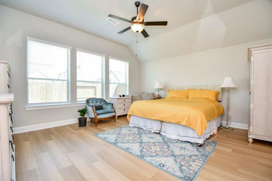 Bright and spacious bedroom with large windows, a vaulted ceiling, and a ceiling fan. Features hardwood floors, a cozy bed with yellow bedding, elegant furniture, and a decorative area rug. Ideal for comfort and relaxation.