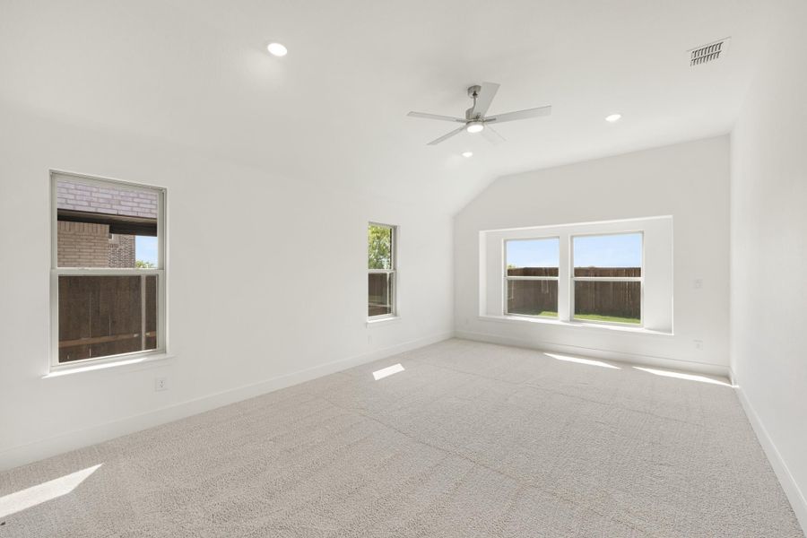 Empty bedroom with vaulted ceiling, ceiling fan, carpet flooring, and three windows with backyard views