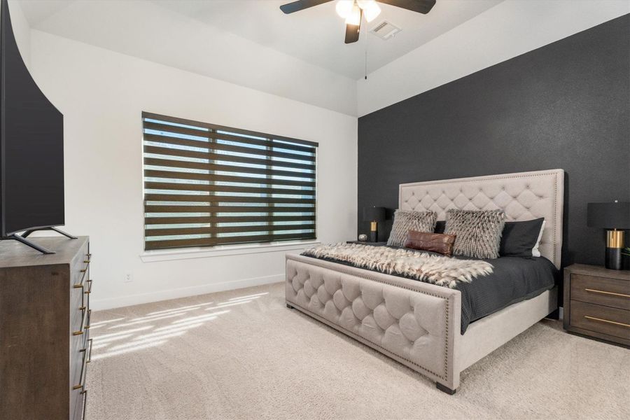 This bedroom features a stylish, modern design with a dark accent wall and a tufted bed. It includes a ceiling fan, patterned window blinds, and a large TV on a dresser, providing a cozy and functional space.