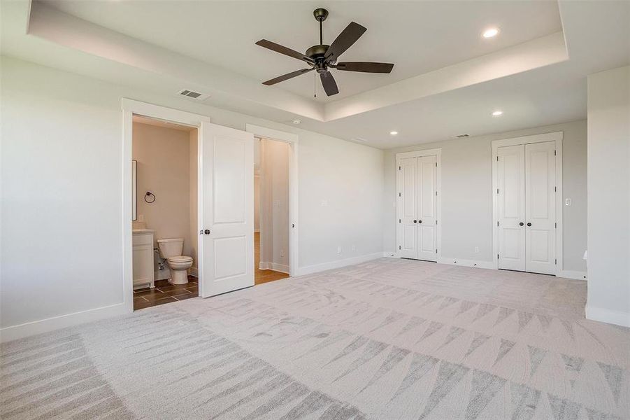 Unfurnished bedroom with ceiling fan, multiple closets, connected bathroom, a tray ceiling, and light colored carpet