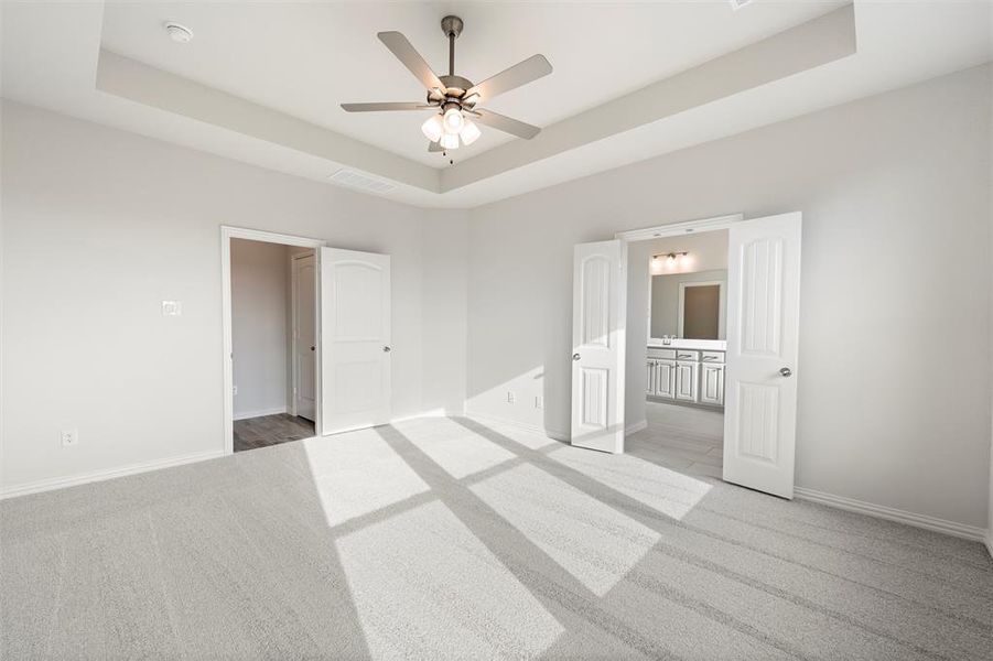 Unfurnished bedroom with a tray ceiling, carpet flooring, ensuite bath, and a ceiling fan