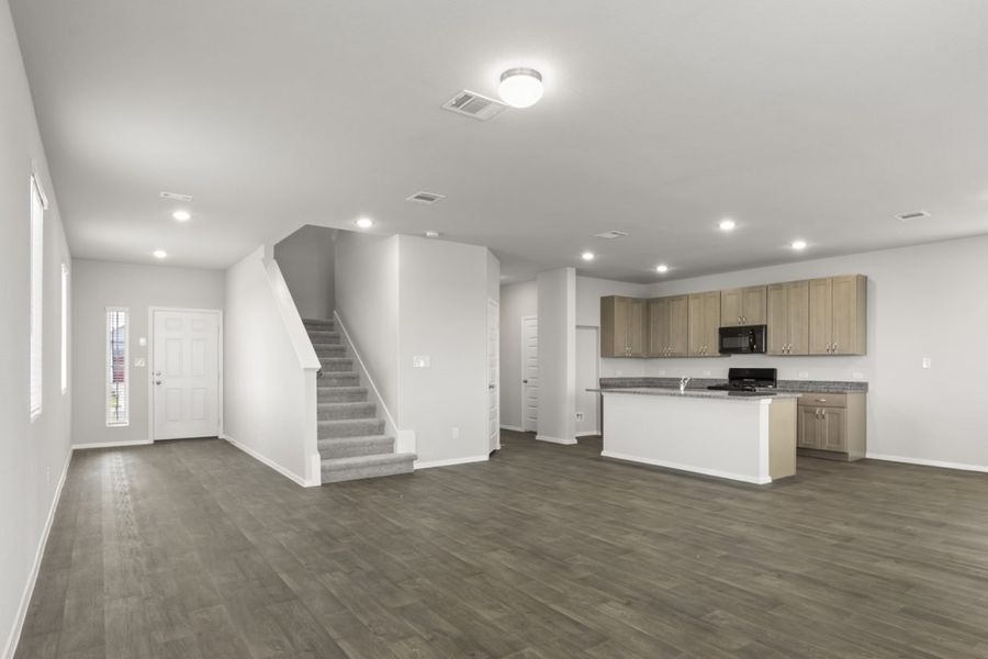 Image of a living room with dark vinyl flooring and grey walls. A front door entry way is on the left next to a carpeted staircase leading upstairs, and a L-shaped kitchen in the corner
