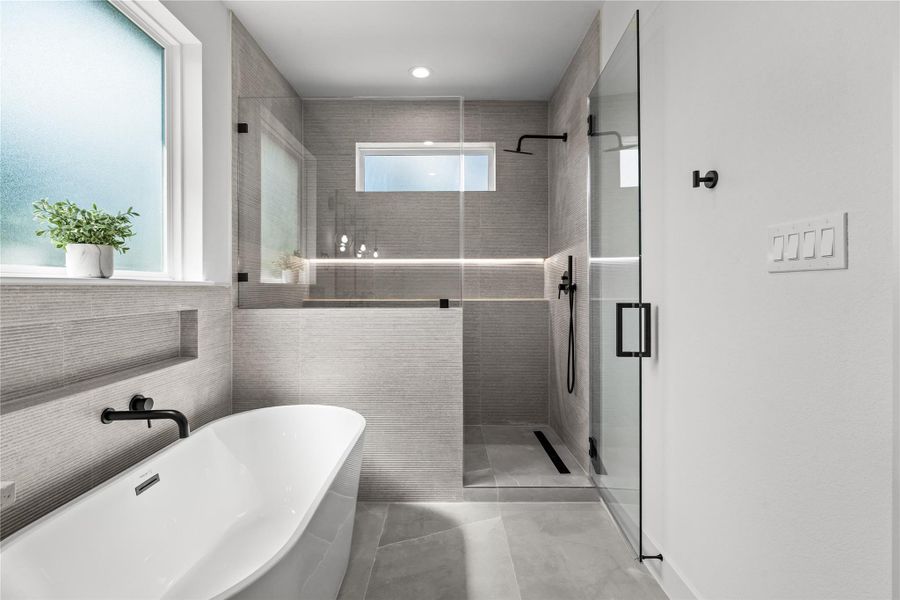 The primary shower features a seamless glass enclosure and neutral floor-to-ceiling tiling. It includes a modern black rainfall showerhead, a built-in tile niche, and a linear drain, sitting right next to the freestanding soaking tub under a large frosted window to create a clean, spa-like wet room.