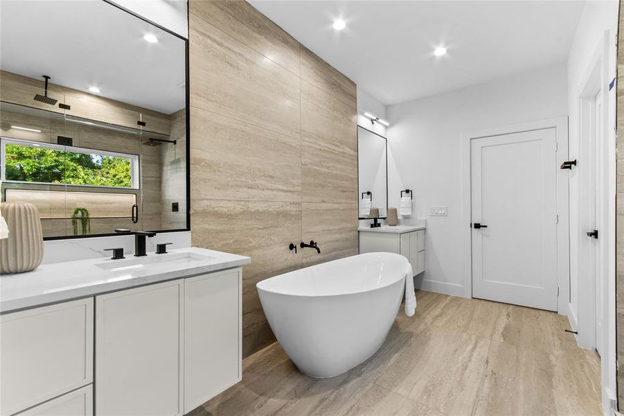 Contemporary bathroom featuring a freestanding soaking tub, dual vanities with white cabinetry and integrated sinks, a walk-in shower with a rain shower head, and floor-to-ceiling textured tile accents Contemporary bathroom featuring a freestanding soaking tub, dual vanities with white cabinetry and integrated sinks, a walk-in shower with a rain shower head, and floor-to-ceiling textured tile accents