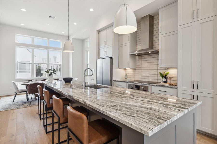 This modern kitchen features a spacious island with elegant stone countertops and seating for four. It boasts sleek cabinetry, a stainless steel range hood, and built-in appliances. The open layout flows into a bright dining area with large windows, offering plenty of natural light.