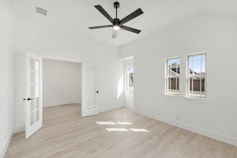 Empty bedroom with light wood floors, black ceiling fan, French door, and multiple windows