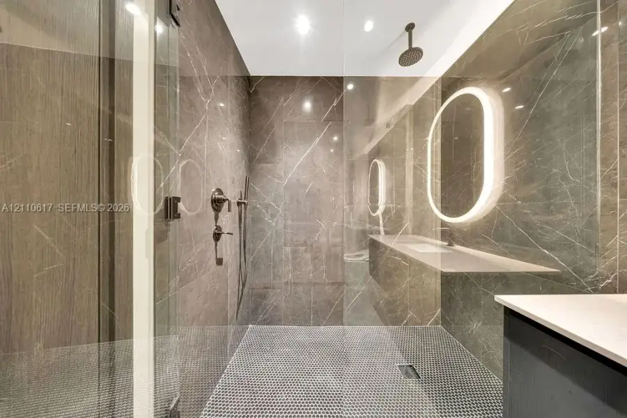 Walk-in shower featuring floor-to-ceiling stone-finish tile, a rainfall showerhead, a frameless glass enclosure, a penny-tile floor, and an integrated illuminated oval mirror Walk-in shower featuring floor-to-ceiling stone-finish tile, a rainfall showerhead, a frameless glass enclosure, a penny-tile floor, and an integrated illuminated oval mirror