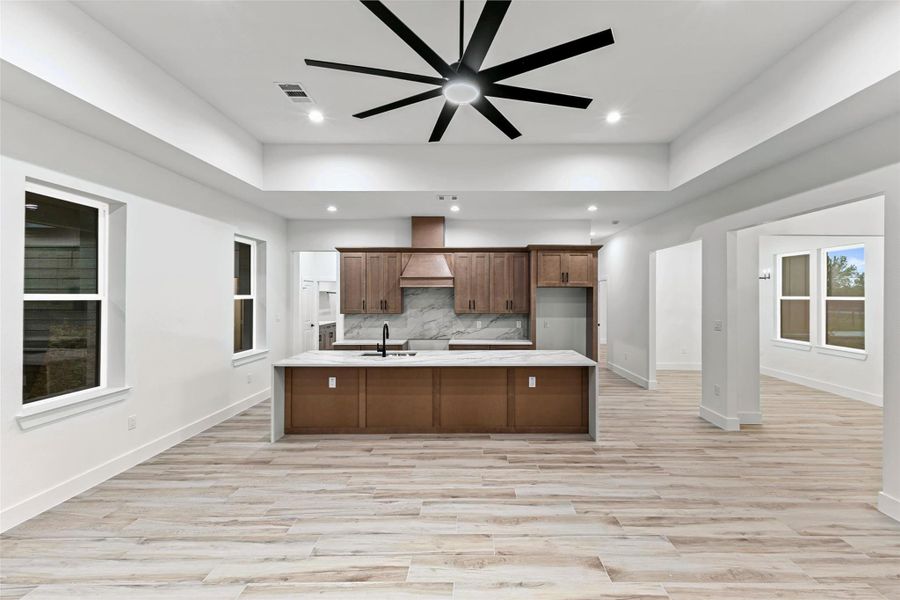 This photo showcases a modern, open-concept kitchen and living area with sleek wood flooring, a large kitchen island, and stylish cabinetry. There's ample natural light from multiple windows, and a striking ceiling fan adds a contemporary touch. This photo showcases a modern, open-concept kitchen and living area with sleek wood flooring, a large kitchen island, and stylish cabinetry. There's ample natural light from multiple windows, and a striking ceiling fan adds a contemporary touch.