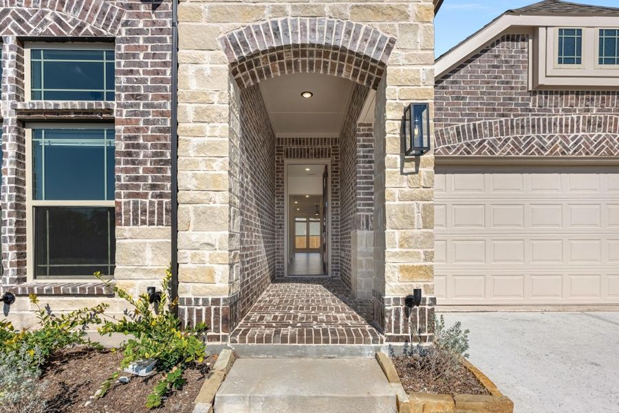 Brick and stone home entrance with arched covered entryway, wall sconce, and two-car garage visible to the right. Brick and stone home entrance with arched covered entryway, wall sconce, and two-car garage visible to the right.