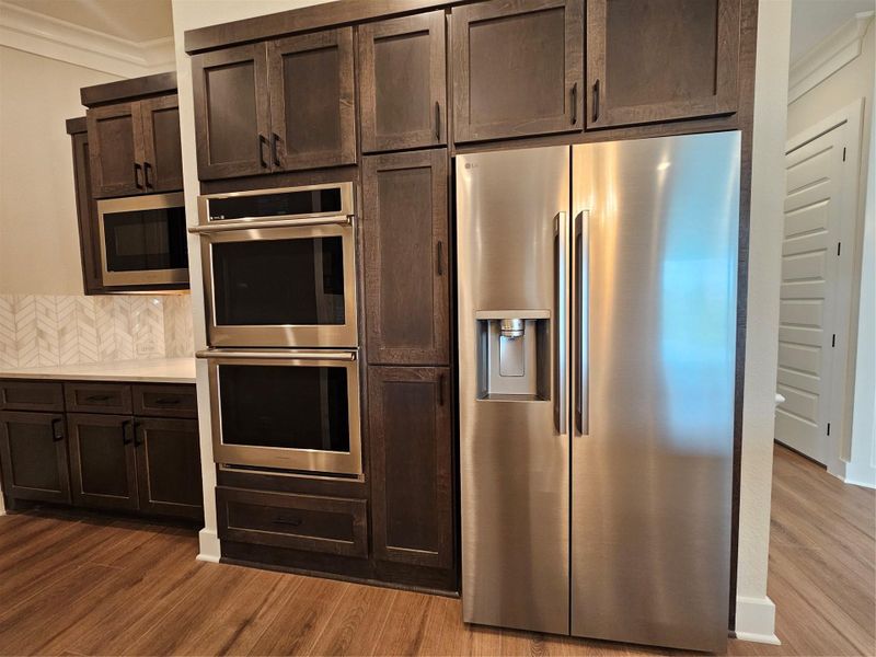 This modern kitchen features dark wood cabinetry, a sleek stainless steel refrigerator with a water dispenser, and a double wall oven. The space is complemented by elegant herringbone backsplash tiles and warm wood flooring. This modern kitchen features dark wood cabinetry, a sleek stainless steel refrigerator with a water dispenser, and a double wall oven. The space is complemented by elegant herringbone backsplash tiles and warm wood flooring.