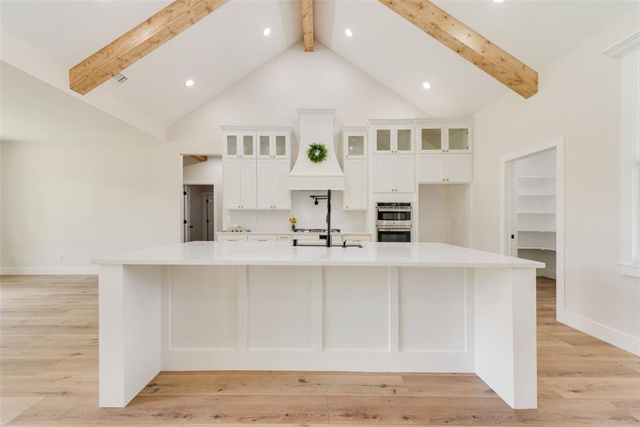 Gourmet kitchen featuring a large central island with white paneling, vaulted ceiling with exposed wood beams, white shaker cabinetry, integrated double ovens, and wood-finish flooring