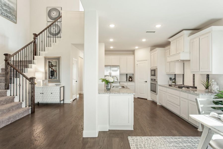 Open-concept kitchen with white cabinets, granite countertops, and dark hardwood floors, adjacent to a staircase with iron balusters.