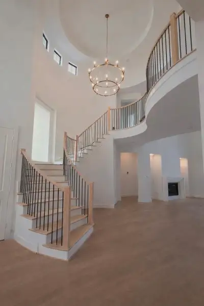 Stunning entryway showcases a grand foyer with an elegant curved staircase featuring light wood handrails and sleek black iron balusters. Also showcasing a floating study, the staircase’s graceful curve, creating a dramatic architectural statement. Abundant natural light pours in through a wall of tall, rectangular windows, highlighting the spacious open floor plan. Fresh white walls, soaring ceilings, and a large dome-style architectural detail above the entry add to the luxurio