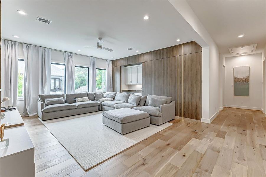 This spacious living area features a large sectional sofa, wood flooring, and ample natural light from four large windows with white curtains. A sleek wood panel wall with custom LED lighting and and built-in storage adds a modern touch, and a ceiling fan enhances comfort.